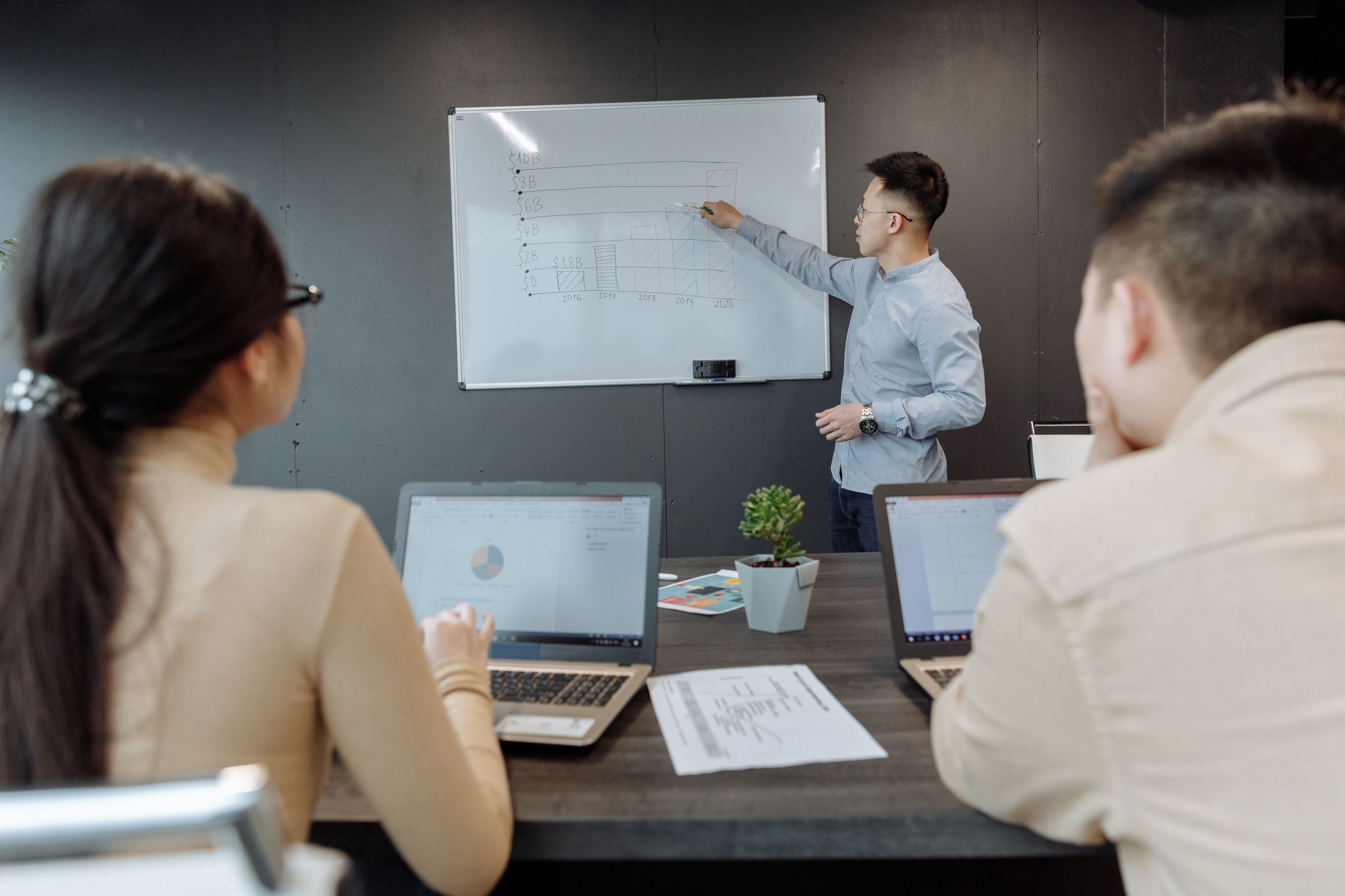 Un formateur explique un graphique au tableau devant un petit groupe équipé d’ordinateurs portables